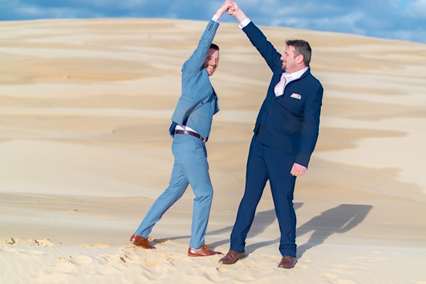 Wide portrait of Tina and Stephen on the Anna Bay sand dunes overlooking the Pacific Ocean at Port Stephens — the vast dune landscape and ocean horizon in spring